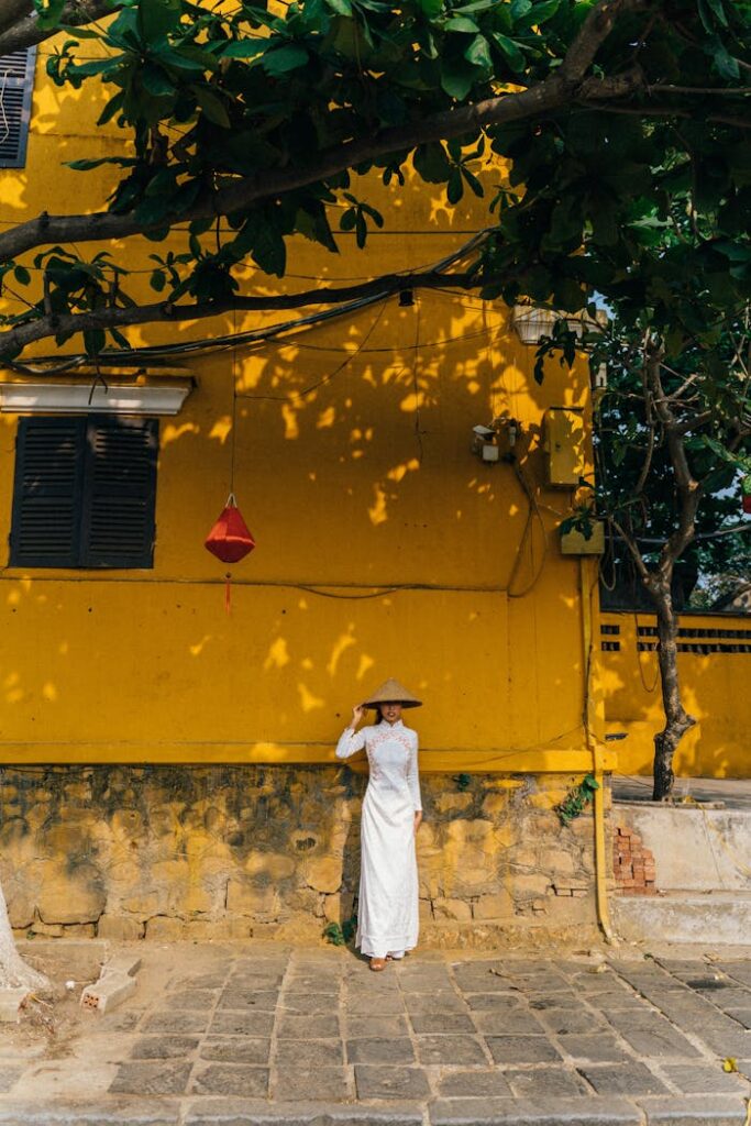 pexels-photo-7216936 A woman in a white Ao Dai stands by a vibrant yellow wall in Hoi An, Vietnam, showcasing traditional culture.